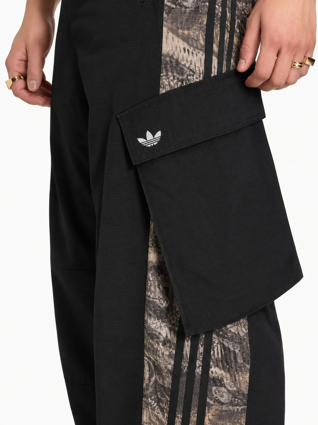Pantalón cargo Adidas negro puesto, detalle del bolsillo cargo con el trébol bordado sobre la franja estampada con las tres rayas