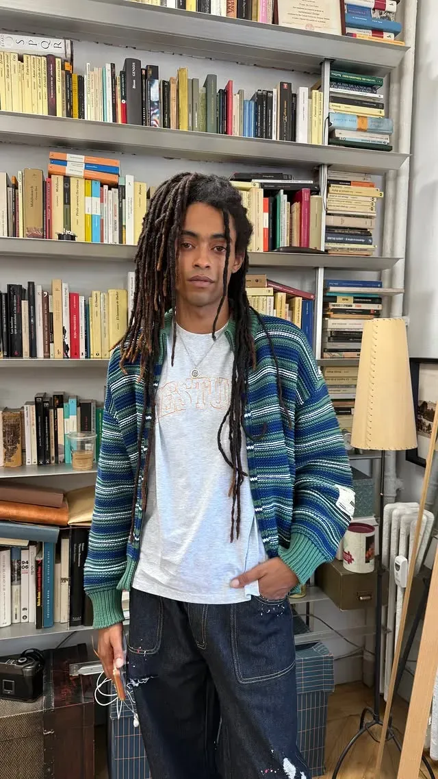 Chico con rastas, cárdigan de rayas azules y verdes y camiseta gris de Eme Studios, delante de una estantería de libros