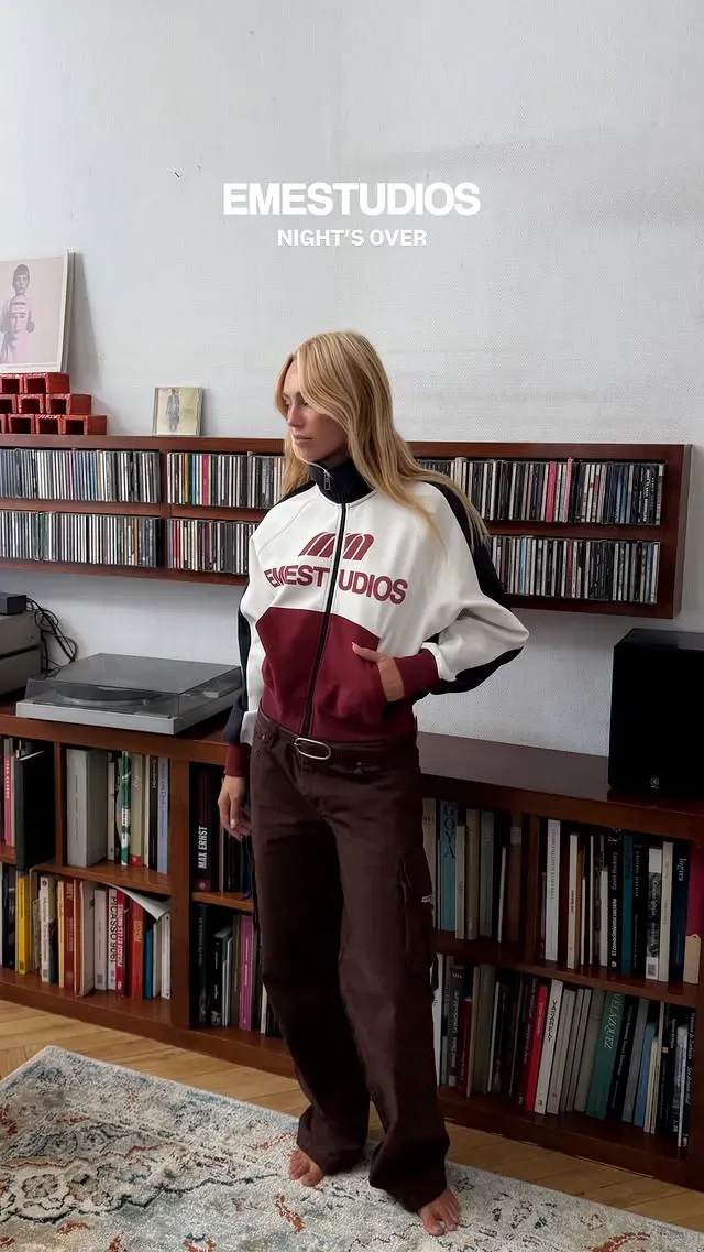 Chica rubia con chaqueta de chándal blanca y granate de Eme Studios y pantalón cargo marrón, en un salón con discos