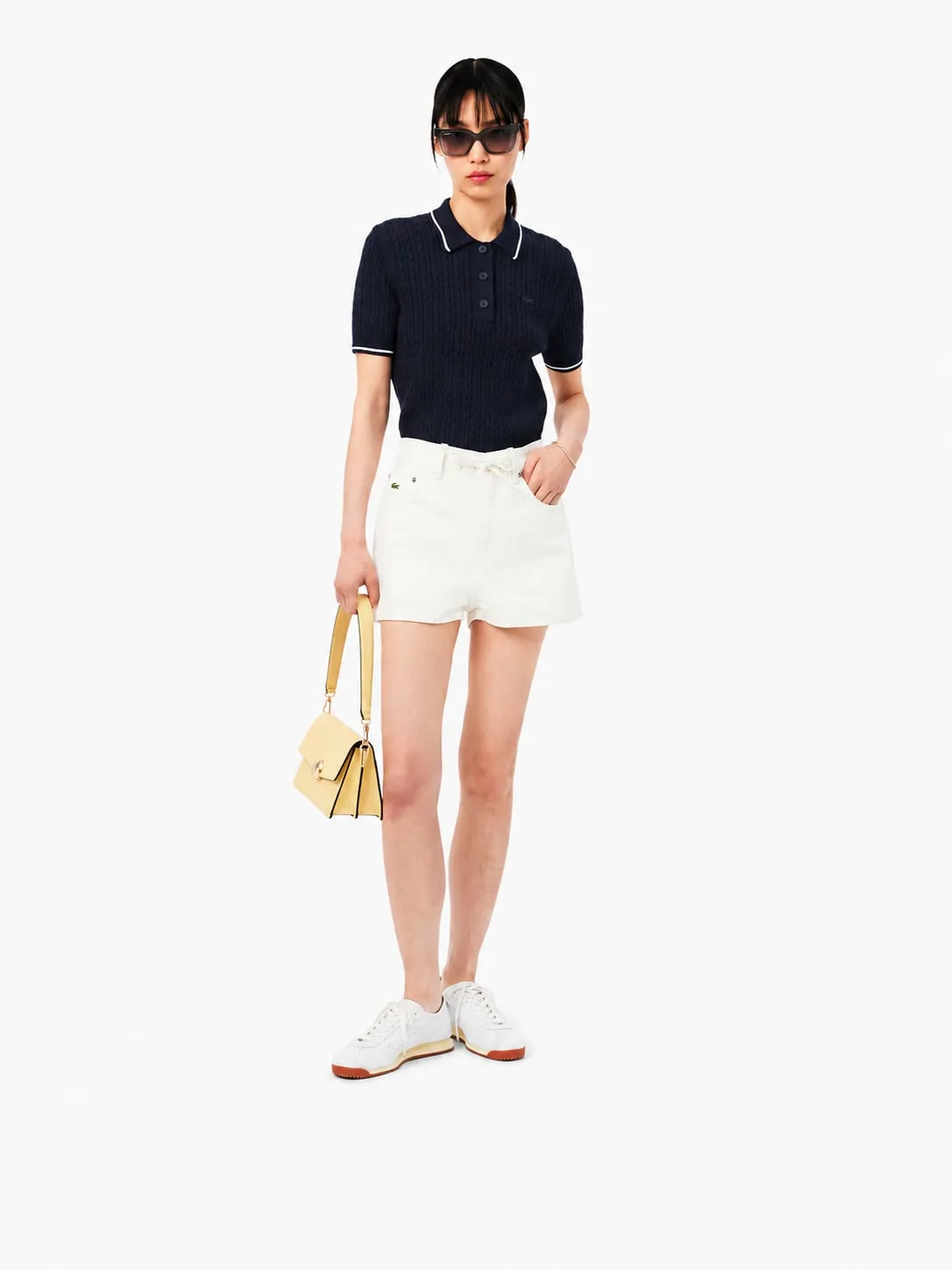 Polo Lacoste de punto trenzado azul marino puesto, plano entero de una modelo con shorts vaqueros blancos y tenis blancos
