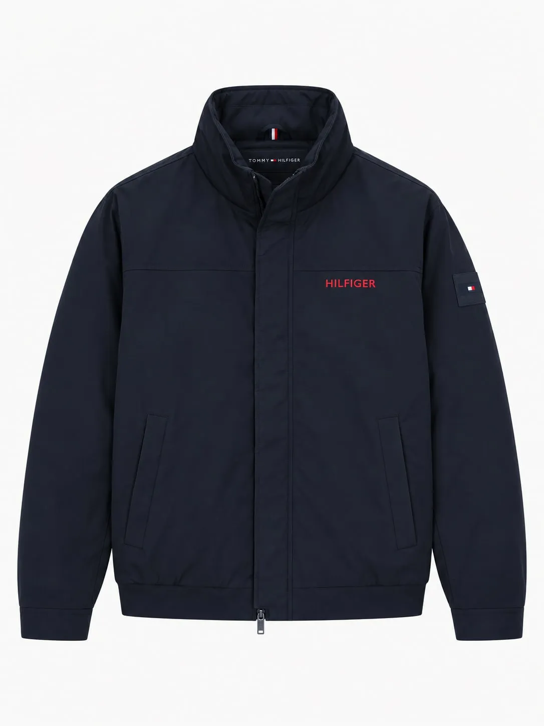 Chaqueta Tommy Hilfiger azul marino, vista frontal de la prenda sola con el cuello alto cerrado, cremallera con tapeta, Hilfiger bordado en rojo en el pecho y la bandera en la manga